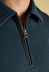 Tumši tēzzili polo krekls ar rāvējslēdzēju ap kaklu, ar strukturētu audumu un kontrastējošu zelta rāvējslēdzēja detaļu.