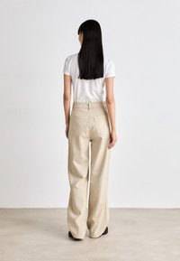 Beige vide bukser med glat tekstur og mellemhøj talje. Parret med en simpel hvid t-shirt, set fra ryggen.