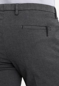 Kalhoty s texturou z uhlíkové barvy se slim fit střihem, disponující zadní kapsou s jemným černým akcentem. Materiál vykazuje jemný tkaný vzor.