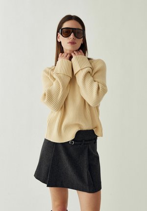 Femme portant un pull beige côtelé oversize, une jupe gris foncé avec ceinture, et de grandes lunettes de soleil foncées, posant devant un fond clair uni.