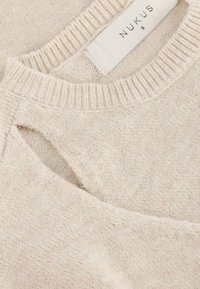 Lichtbeige gebreide trui met een ronde halslijn, uitsnijding op de schouder en een geweven label met de tekst "NUKUS" maat S.