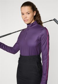 Vijolična polo majica z dolgimi rokavi in visokim ovratnikom, ki ima kontrastne roza črte po rokavih, kombinirana s prilegajočo se črno krilo. Golf palica počiva na rami.