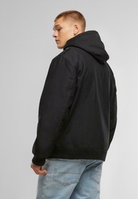 Black hooded jacket with a smooth fabric, elastic cuffs, and a relaxed fit, paired with light blue denim jeans.