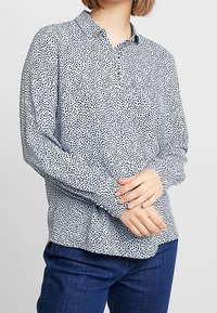 Langärmlige Bluse aus hellblauem Stoff mit schwarzen Punkten, mit einem Knopfkragen und einem lässigen Schnitt. Getragen mit dunkelblauen Jeans.