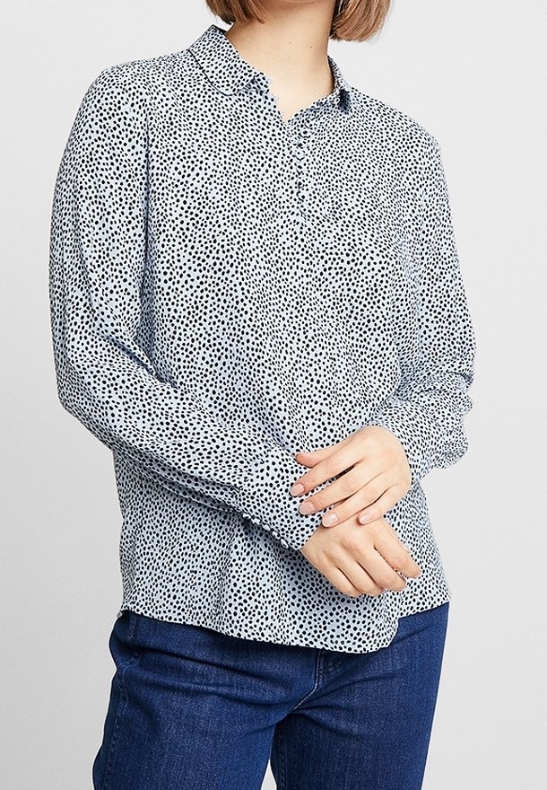 Langärmlige Bluse aus hellblauem Stoff mit schwarzen Punkten, mit einem Knopfkragen und einem lässigen Schnitt. Getragen mit dunkelblauen Jeans.