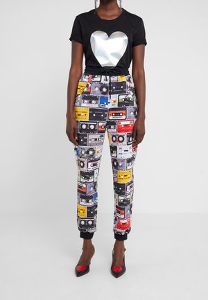 Osoba ubrana w czarną koszulkę z dużym srebrnym sercem oraz kolorowe spodnie dresowe pokryte wzorami kaset magnetofonowych, do tego czarne szpilki z czerwonymi sercami.