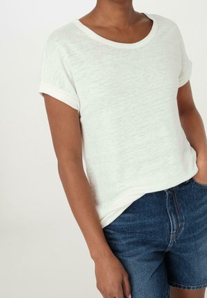 Persoon die een wit gestructureerd T-shirt met korte mouwen draagt, ingestopt in een hooggetailleerde blauwe denimshort, met één hand in de zak.