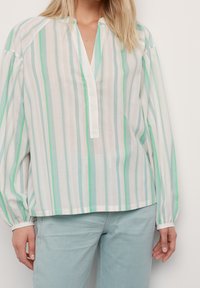 Blusa leggera a righe verde e bianco con parziale apertura a bottoni, maniche lunghe a sbuffo e vestibilità rilassata. Abbinata a pantaloni azzurri chiari.