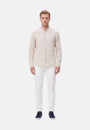 Uomo in piedi di fronte che indossa una camicia beige con bottoni e collo alla mandarino, pantaloni bianchi e scarpe da ginnastica blu navy su sfondo bianco.