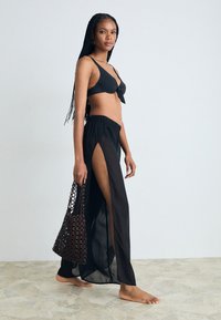 Crni bikini s detaljem vezanja sprijeda uparen s prozirnom crnom suknjom s visokim prorezom do bedara. Model drži ručno rađenu torbu, bosa na kamenitoj površini.