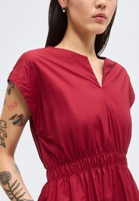 Γυναίκα με κοντομάνικο, βαθύ κόκκινο φόρεμα με ζωνάκι στη μέση και V-λαιμόκοψη, που δείχνει τατουάζ στο αριστερό της μπράτσο και σκουλαρίκι με πέρλα.