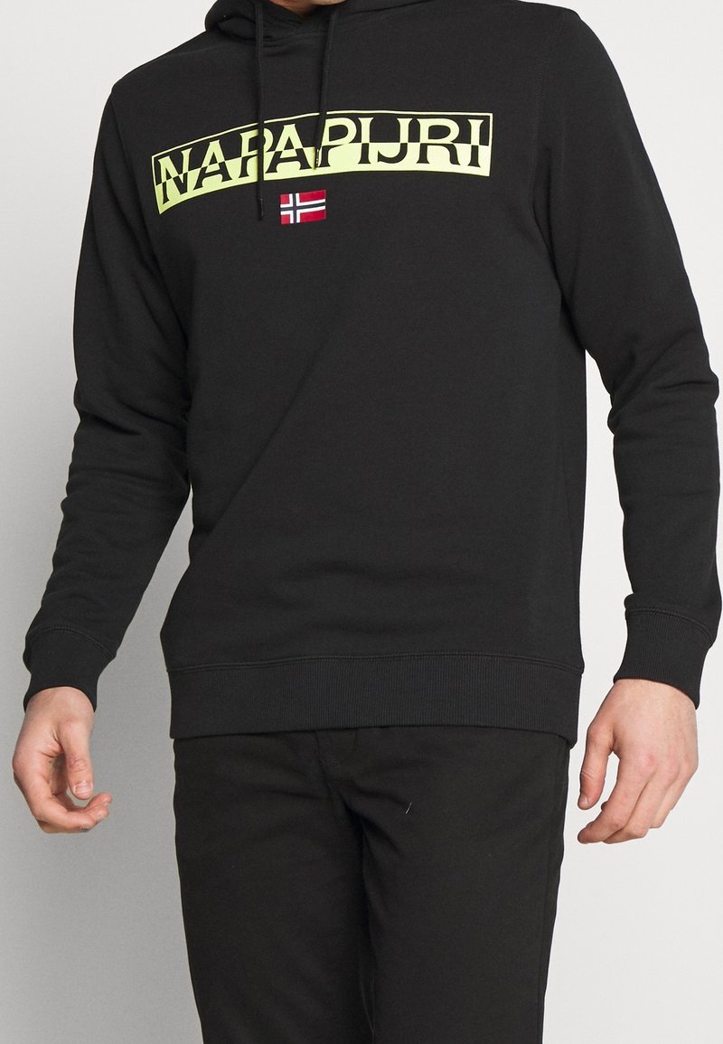Homme portant un sweat à capuche noir avec le texte jaune "NAPAPIJRI" et un petit drapeau norvégien rouge et blanc sur la poitrine.