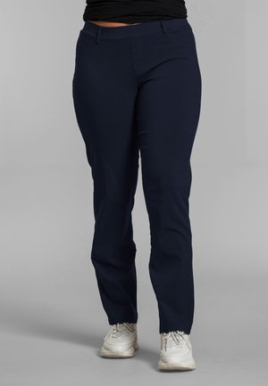 Person iført marineblå bukser med lige ben og hvide sneakers, stående mod en ensfarvet grå baggrund.