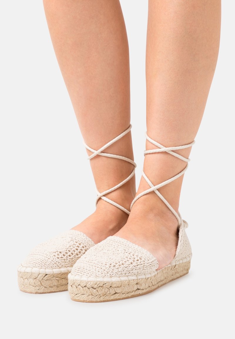 Pieds portant des espadrilles plates tissées beige avec lacets à enrouler autour de la cheville et semelles compensées en jute sur un fond uni.
