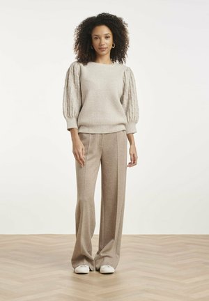 Vrouw met krullend haar gedragen beige gestructureerde trui met pofmouwen, beige wijde broek, witte sneakers, staand op een lichte houten vloer.