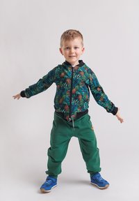 Kurtka bomber z zamkiem błyskawicznym w ciemnozielonym kolorze z wzorem szyszek i liści, zestawiona z luźnymi zielonymi spodniami i niebieskimi trampkami.