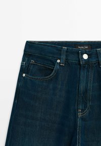 Jean en denim bleu foncé avec poches avant, passants pour ceinture, fermeture par bouton et coutures visibles, étiqueté Massimo Dutti.
