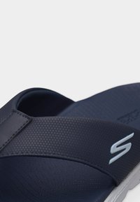 Κοντινό πλάνο ενός σαγιοναριού σε σκούρο μπλε με υφασμένους ιμάντες και λευκό λογότυπο "S" στο πλευρό, πάνω σε λευκό φόντο.