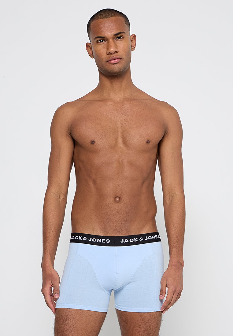 Hombre joven de pie, vestido con bóxer azul claro con cinturilla negra de "Jack & Jones", frente a un fondo blanco liso.