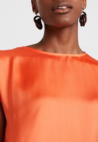 Top in raso arancione con scollo rotondo e maniche corte. Notevoli orecchini circolari scuri accentuano il décolleté. Telaio liscio.