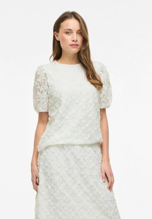 Femme portant un haut à manches ballon en dentelle florale blanche et une jupe assortie, debout devant un fond blanc uni.