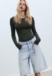 Grünes Langarmshirt, kombiniert mit hellblauen Denim-Shorts, die ein weites Bein haben und einen dekorativen, mit Perlen besetzten Schlüsselanhänger am Bund tragen.