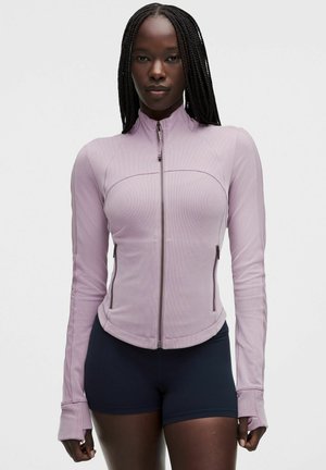 Kvinde med langt flet hår iført en tætsiddende lavendelfarvet lynlåstrøje og sorte shorts mod en enkel, lys baggrund.