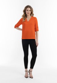 Orange, T-shirt med V-hals og opbrettede ærmer, iført med sorte stramme bukser og sorte stiletter. Glat stof med en afslappet pasform.