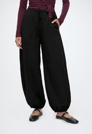 Femme portant un pantalon noir ample avec des chevilles élastiques, un haut bordeaux à manches longues et des chaussures plates noires à brides.
