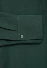 Detail zelené košile s límečkem, která má složený manžetový rukáv se střapcovým knoflíkem, hladkou texturu látky a jemné plizé na rukávu.