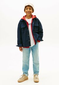 Granatowa dżinsowa kurtka noszona na czerwonym hoodie, białej koszulce, jasnoniebieskich dżinsach i beżowych sneakersach. Na głowie czapka z dzianiny dopełnia stylizację.