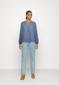 Blusa azul com bolinhas brancas, com decote em V e botão, apresentando mangas longas e um corte solto, combinada com jeans azuis claros e botas de salto castanhas.