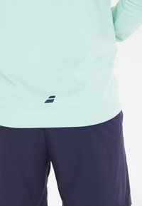 Lys mintgrøn langærmet skjorte med en tætsiddende pasform og en mørk marineblå kant ved kanten. Kombineret med marineblå shorts, glat tekstur.