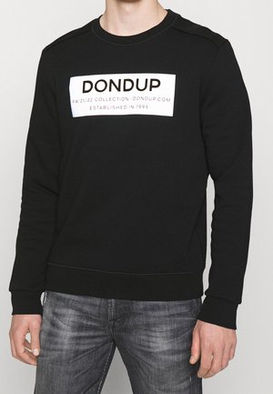 Mężczyzna w czarnej bluzie z nadrukiem "DONDUP" na piersi w białym bloku, w zestawie z wyblakłymi szarymi dżinsami, stojący na jednolitym tle.