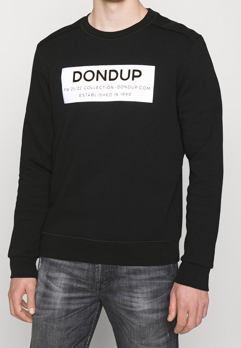 Homme portant un sweat noir avec "DONDUP" imprimé sur la poitrine en lettres blanches, assorti à un jean gris délavé, debout devant un fond uni.