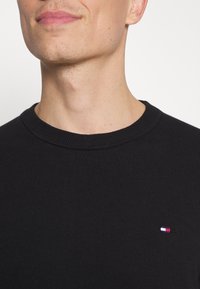 Schwarzer Baumwollstrickpullover mit geripptem Rundhalsausschnitt und einem kleinen, mehrfarbigen Logo auf der Brust.