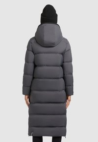 Abrigo acolchado gris con capucha, diseño acolchado, mangas largas y silueta hasta la rodilla. Se lleva con un gorro de punto negro.