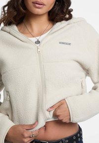 Giacca in pile color crema con chiusura a zip, tasche frontali e una piccola etichetta nera "Guess Jeans". Tessuto testurizzato e design aderente.