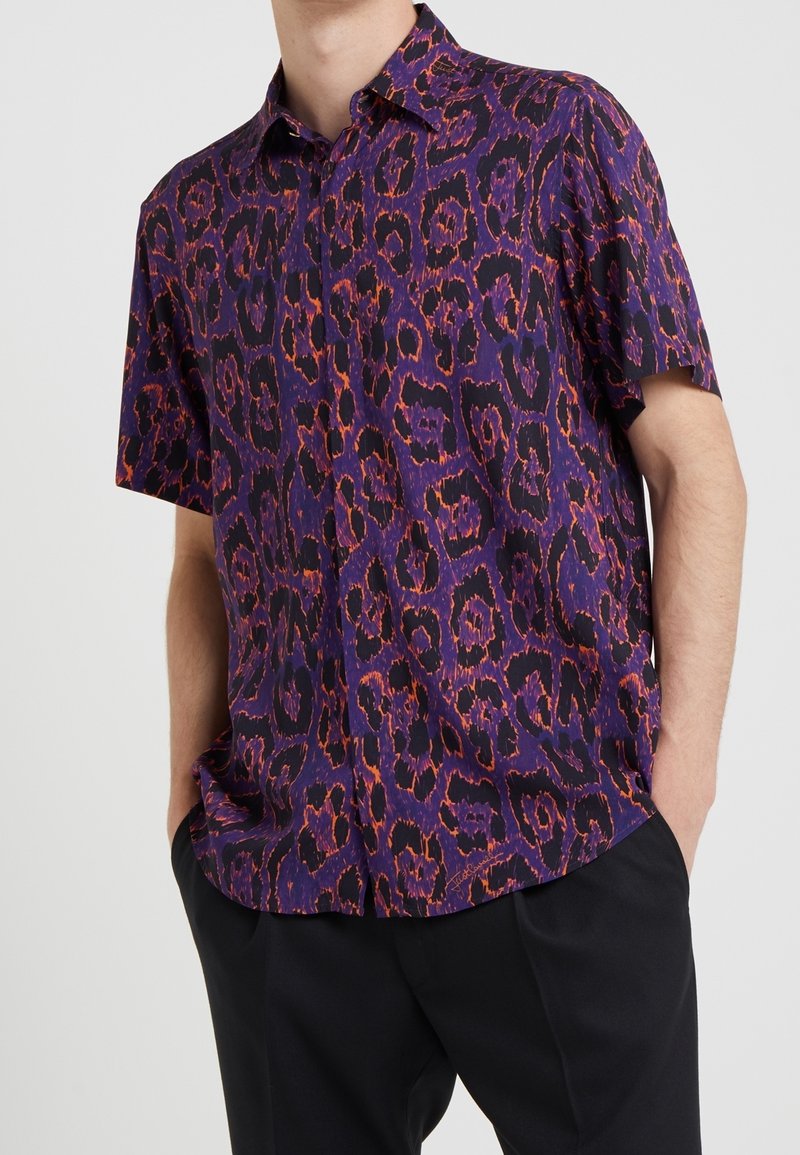 Homme portant une chemise à manches courtes boutonnée avec un imprimé léopard abstrait violet et orange, associée à un pantalon noir, mains dans les poches.