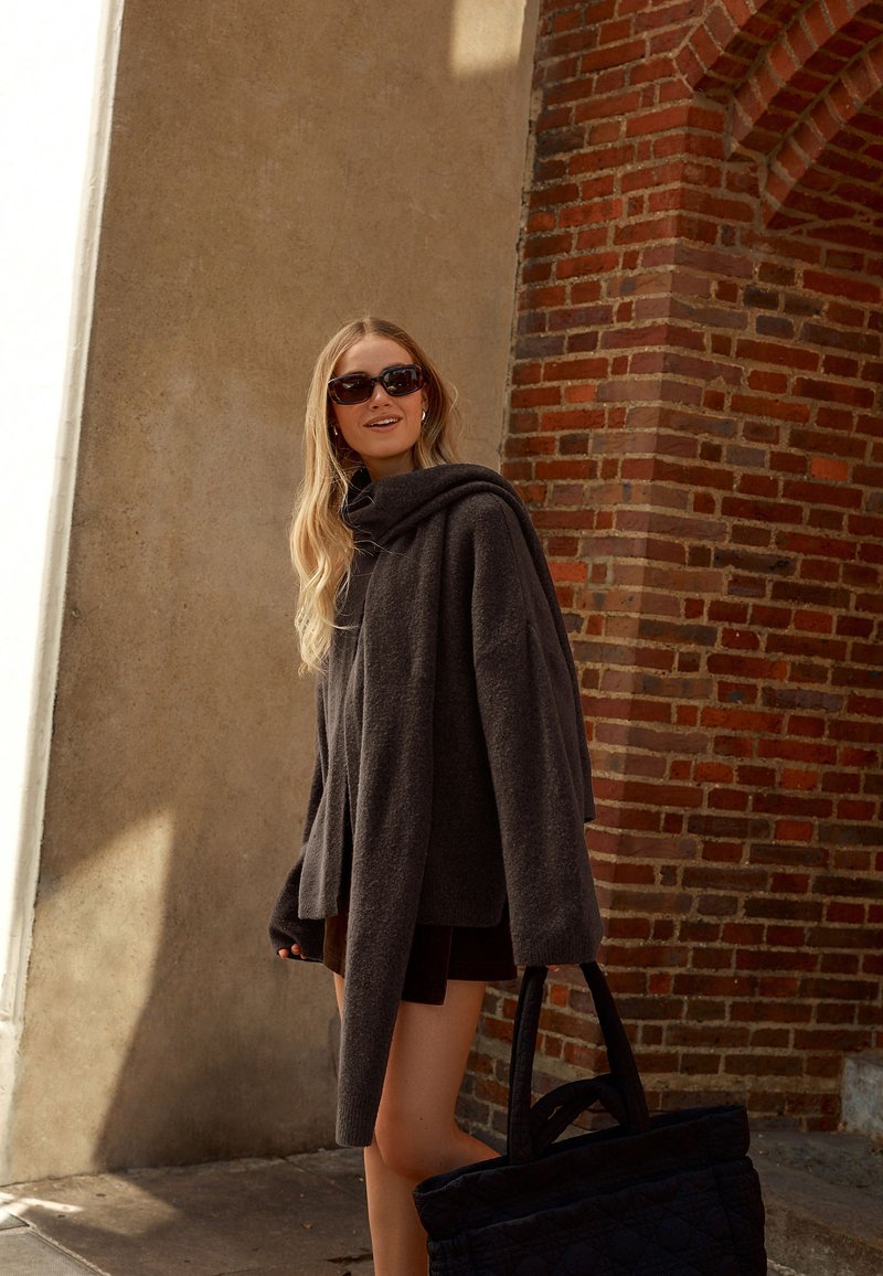 Jeune femme aux longs cheveux blonds porte un pull oversize sombre, des lunettes de soleil, tenant un grand sac noir, debout près d'un mur en briques rouges et un béton ombragé.