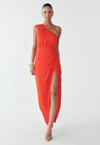 Model in een levendige oranje een-schouderjurk met een hoge split tot aan de bovenbenen, gestyled met een armband en minimale make-up, staat tegen een effen achtergrond.