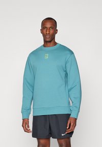 Sudadera de color verde azulado con mangas largas, cuello redondo y un pequeño logo amarillo en el centro. Llevada con pantalones cortos negros que tienen un swoosh blanco.