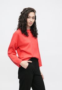 Spilgti koraļļu džemperis ar apaļu kakla izgriezumu un ribotām aprocēm, kombinēts ar melnām biksēm. Gluda tekstūra, brīvs piegriezums un garas piedurknes.