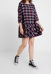 Femme portant une robe à carreaux bleu marine, rouge et blanc avec des manches trois-quarts et des baskets montantes noires, marchant devant un fond blanc.