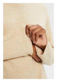 Beige gebreide trui met geribbelde manchetten. Zachte textuur, iets chunky ontwerp. Hand die voorzichtig de manchette vasthoudt, waardoor de structuur van het kledingstuk wordt benadrukt.