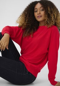 Punainen neulepaita, jossa on rento malli, roikkuvat olkapäät ja ribbaatut hihansuut, yhdistettynä tummiin farkkuihin. Silkkinen pinta, rento muotoilu.