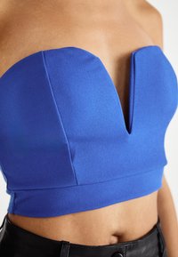 Modrý crop top bez ramienok s hlbokým výstrihom do V, vyrobený z hladkej látky. Obsahuje štruktúrované švy a priliehavý strih, ktorý sa dobre kombinuje s nohavicami s vysokým pásom.