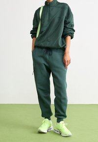 Osoba v tmavě zelené texturované mikině se zipem a odpovídajících teplácích, neonově zelených teniskách, stojící na světle zelené podlaze s rukama v kapse.