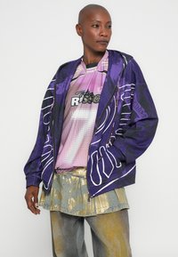 Giacca bomber viola con disegni astratti bianchi, indossata sopra una maglia grafica rosa e una gonna metallica dorata, che mostra una miscela di texture.
