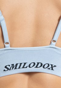 Sujeción deportiva de color azul claro con textura acanalada y texto bordado en negro "SMILODOX" en la banda. Tirantes delgados ajustables y textura suave.